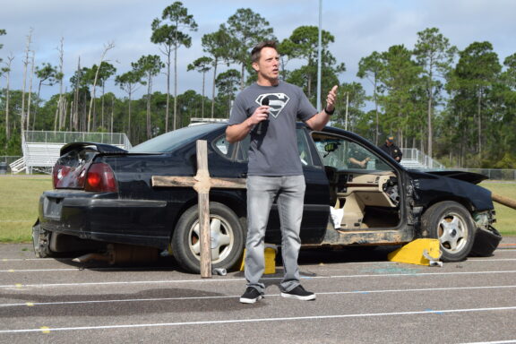 man standing in front of a wrecked car talking on a microphone