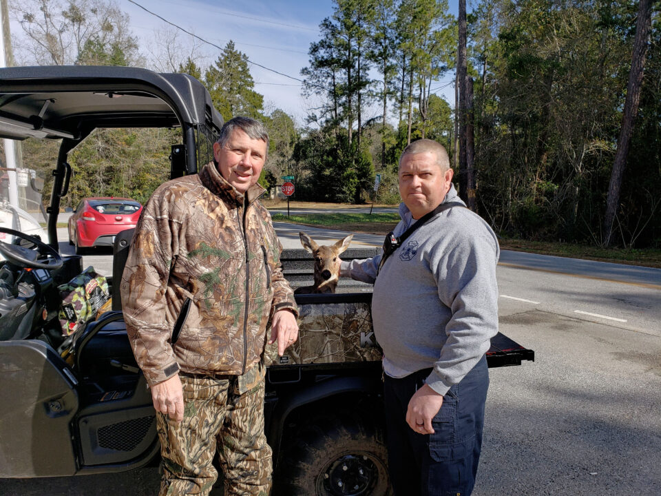 Man in camouflage jacket and camouflage pants and a firefighter in navy pants and a grey pullover standing in front of a camouflage utility vehicle with a young deer inside