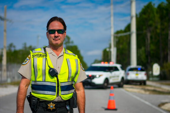 A white male with sunglasses on stands in the roadway wearing a safety vest.,