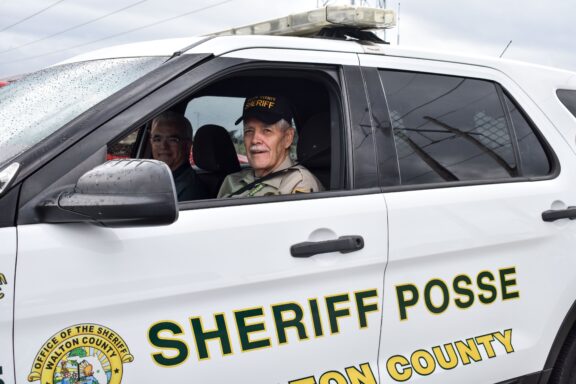A white male with white hair wearing a beige uniform sits in a patrol car marked "Sheriff Posse"