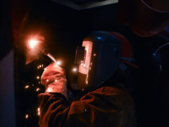 A woman wearing a welding helmet sparks her torch as she welds.