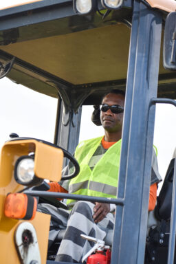 A black male with sunglasses on in an inmate uniform drivers a front-end loader.