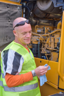 White male with sunglasses on in an inmate uniform checks the oil in an excavator
