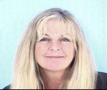 Mug shot of a white woman with blonde hair in her 50's smiling wearing a navy blue jump suit.