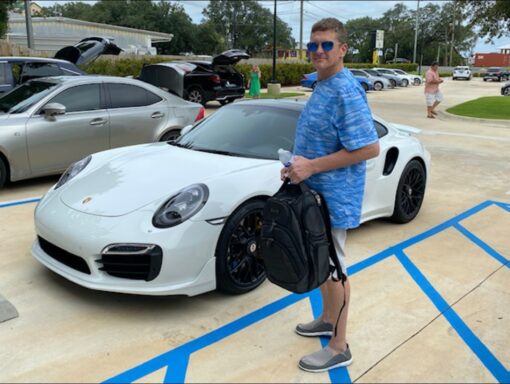 A white male in a blue shirt holding a backpack. In the backround is a white Porsche Turbo.