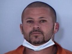 Mug shot of white male with facial hair and a mask hanging below his chin.