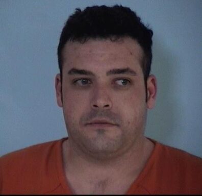 Mug shot of a white male with dark hair wearing an orange jumpsuit.