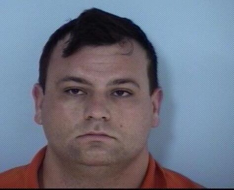 A mug shot of a white male with dark hair and features with an orange jumpsuit on.