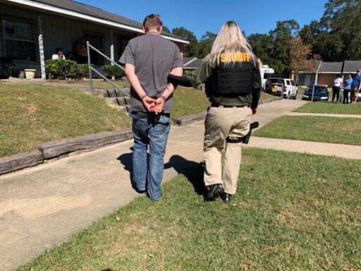 A female deputy with a bullet-proof vest walks a white male in handcuffs along a sidewalk in front of an apartment complex.