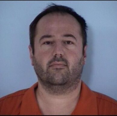 Mug shot of a white male with dark hair and short facial hair wearing an orange jumpsuit