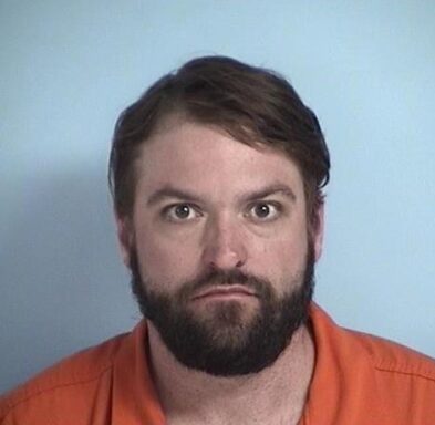 Mug shot of a white male with dark brown facial hair wearing an orange jumpsuit