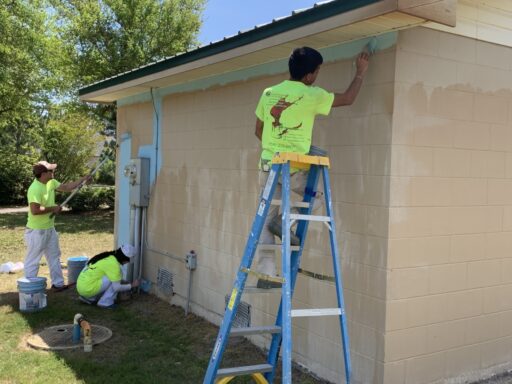 Three individuals with bright yellow shirts painting the side of a building with light blue paint
