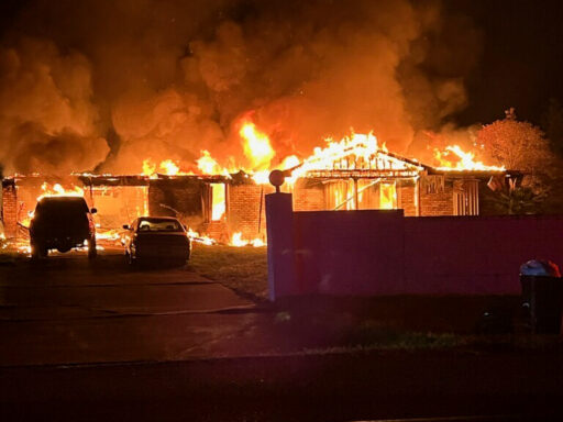 House engulfed in flames with a truck and car parked in the driveway