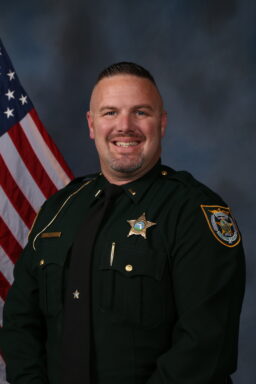 A white male with facial hair in a green deputy sheriff uniform with a five star badge and a gold pen.