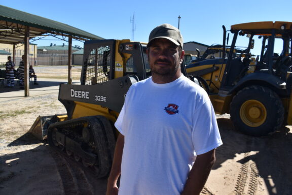 A man in a white t-shirt poses in front of a skid steer