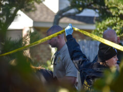 A medical examiner lifts crime scene tape to allow an investigator out of the taped off area.