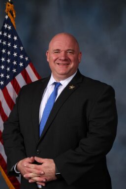 White man with a suit and royal blue tie smiling standing in front of an American flag.