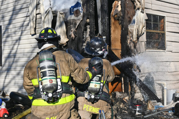 Firefighter holding hose spraying water onto fire damaged home with second firefighter behind her for support