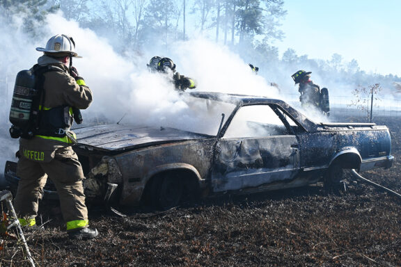 Firefighters spraying water on a car that caught on fire with heavy smoke coming out of the vehicle