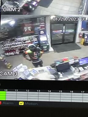 A black male wearing a black hoodie and mask standing in a gas station.