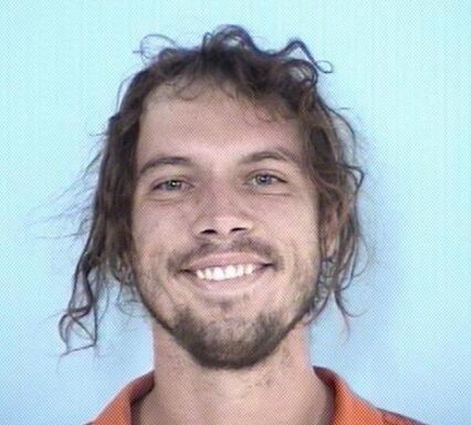 Mug shot of a white male with long brown hair tied back in a bun wearing an orange jumpsuit smiling.
