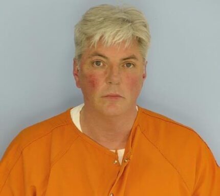 Mug shot of a white male with white hair and a red abrasion on his right cheek wearing an orange jumpsuit.