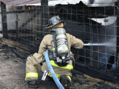 Firefighter wearing black helmet squatting down, spraying water through a wire fence into a barn that caught on fire