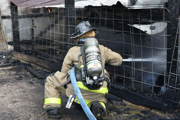 Firefighter wearing black helmet squatting down, spraying water through a wire fence into a barn that caught on fire