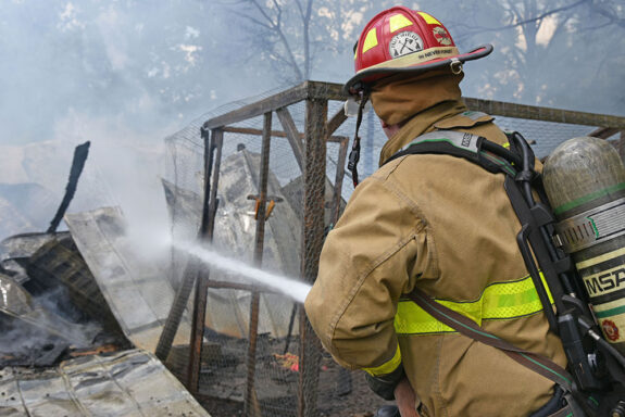 Firefighter wearing red helmet spraying water on a barn that is on fire