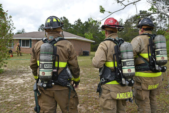 Three firefighters in bunker gear looking at a single-story brick house