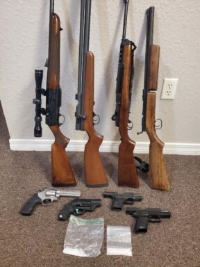Four long guns leaning against a wall with four hand guns on the floor and two clear bags of white substance on the floor.