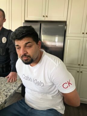 Male with facial hair wearing a Calvin Klein shirt handcuffed with an officer standing behind him