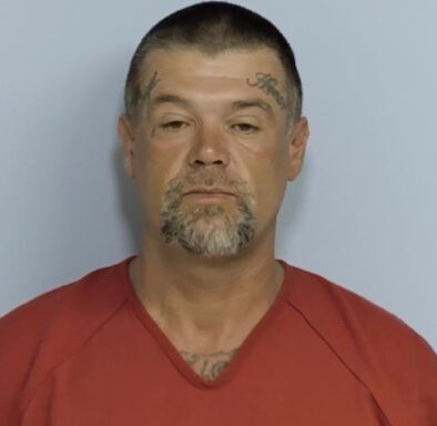 Mug shot of a white male in an orange jumpsuit with facial tattoos on his temples and a white and gray goatee.