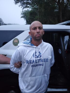 White male in a white "Realtree Fishing" sweatshirt standing in front of a Walton County Sheriff's Office patrol car in handcuffs.