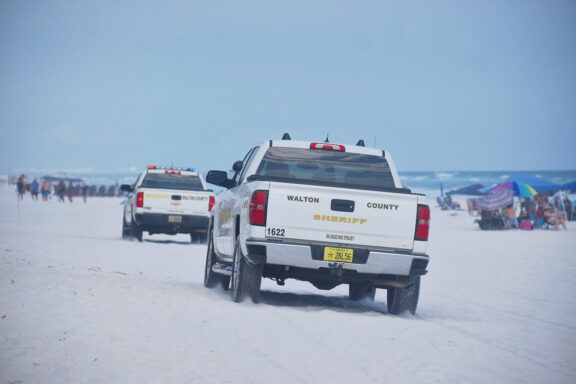 Two white trucks with Walton County Sheriff's Office logos drive down the beach on white sand.