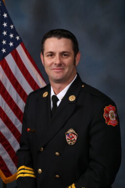 White male with dark brown hair wearing a navy blue fire rescue formal jacket, a white collar shirt, and a navy blue tie