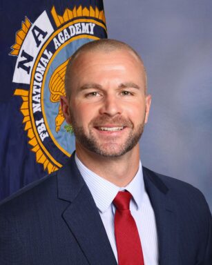 A white male with brown hair and brown facial hair wearing a blue suit with a red tie smiles in front of the FBI National Academy flag against a blue back drop.