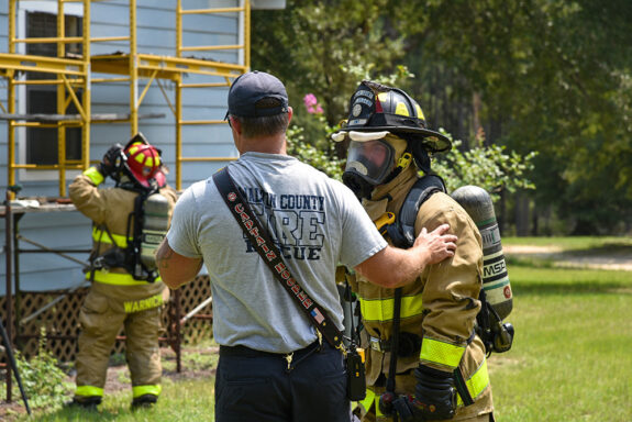 Fire department captain wearing a gray shirt and navy pants standing next to a firefighter in bunker gear