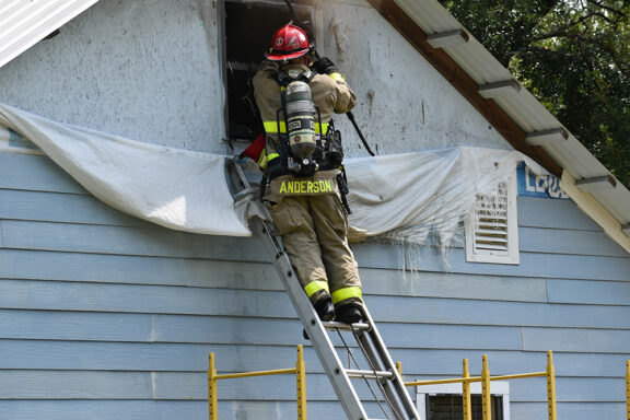 Firefighter wearing red helmet on a ladder outside of a light blue home