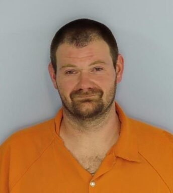 Mug shot of a white male with brown hair and red glossy eyes wearing an orange jumpsuit. Man also has brown facial hair. 