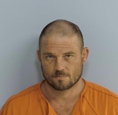 Mug shot of white male with thinning brown hair and brown beard wearing an orange jumpsuit