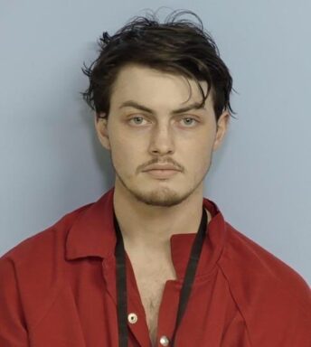 Mug shot of a white male with shaggy dark brown hair and a small goatee wearing an orange jumpsuit with a black lanyard around his neck.