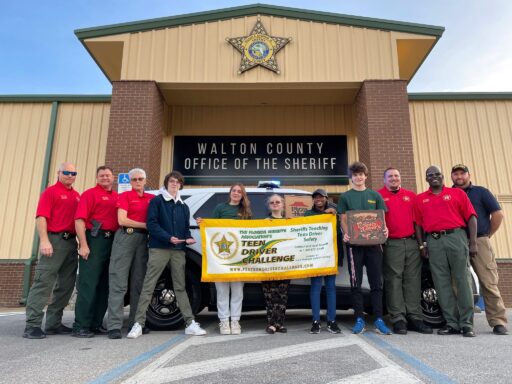 A group of teens standing with instructors wearing red shirts holding up a Teen Driver Challenge banner