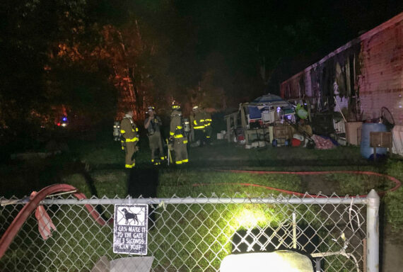 Firefighters in bunker gear standing in yard outside of single-wide mobile home with fire damage