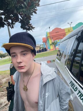 Photo of a white male in handcuffs with a gold chain and a hat on. 