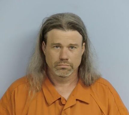 A white male with long blonde/graying hair with a salt and pepper goatee wearing and orange jumpsuit.