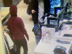 Two males standing at a cashiers counter one is wearing a maroon sweatshirt the other is wearing a black zip-up hoodie.