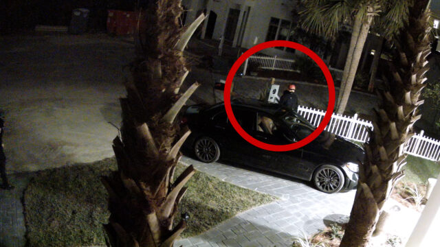 Still shot of a suspect at a dark vehicle wearing an orange hat attempting to burglarize a black vehicle sitting in a drive way.
