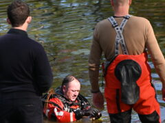 White male wearing navy blue pants and sweatshirt standing beside another man wearing dive gear on the banks of a pond where a diver is waste deep adjusting his dive mask.