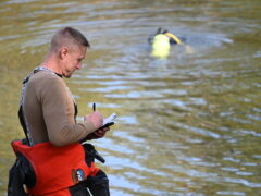 A white male holds a clip board wearing a longsleeve brown shirt standing on the banks of a pond where a diver is in the water.